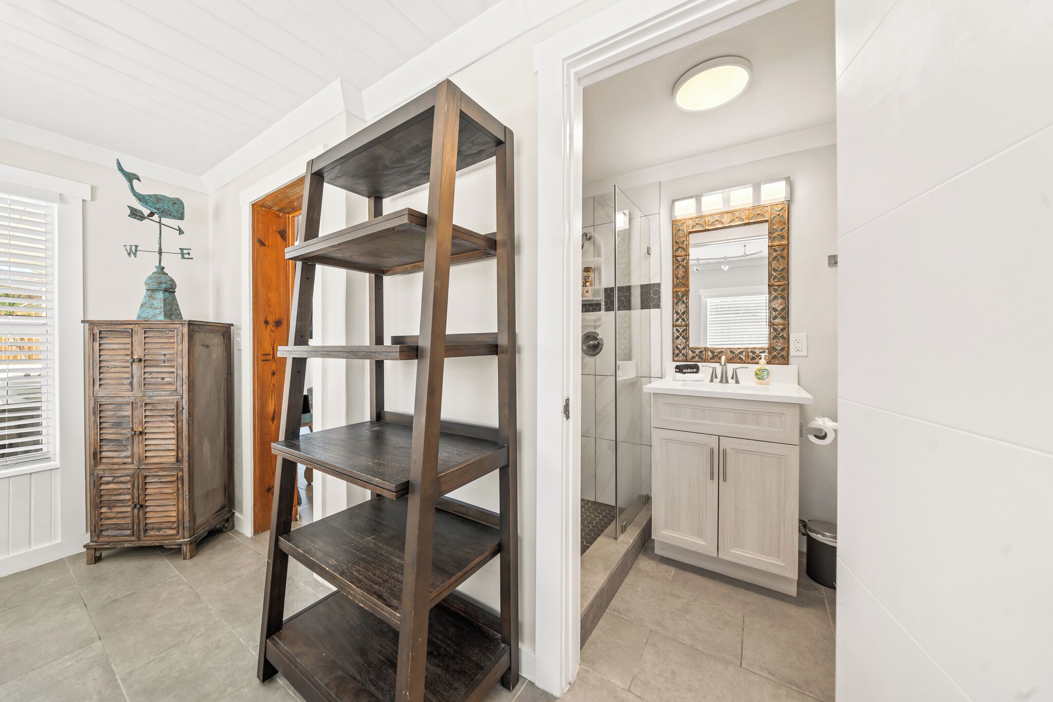 A room with a wooden shelving unit, a cabinet with a decorative top, and a bathroom with a mirror and sink.