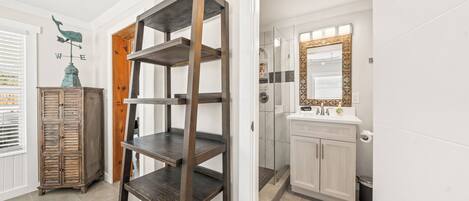 A room with a wooden shelving unit, a cabinet with a decorative top, and a bathroom with a mirror and sink.