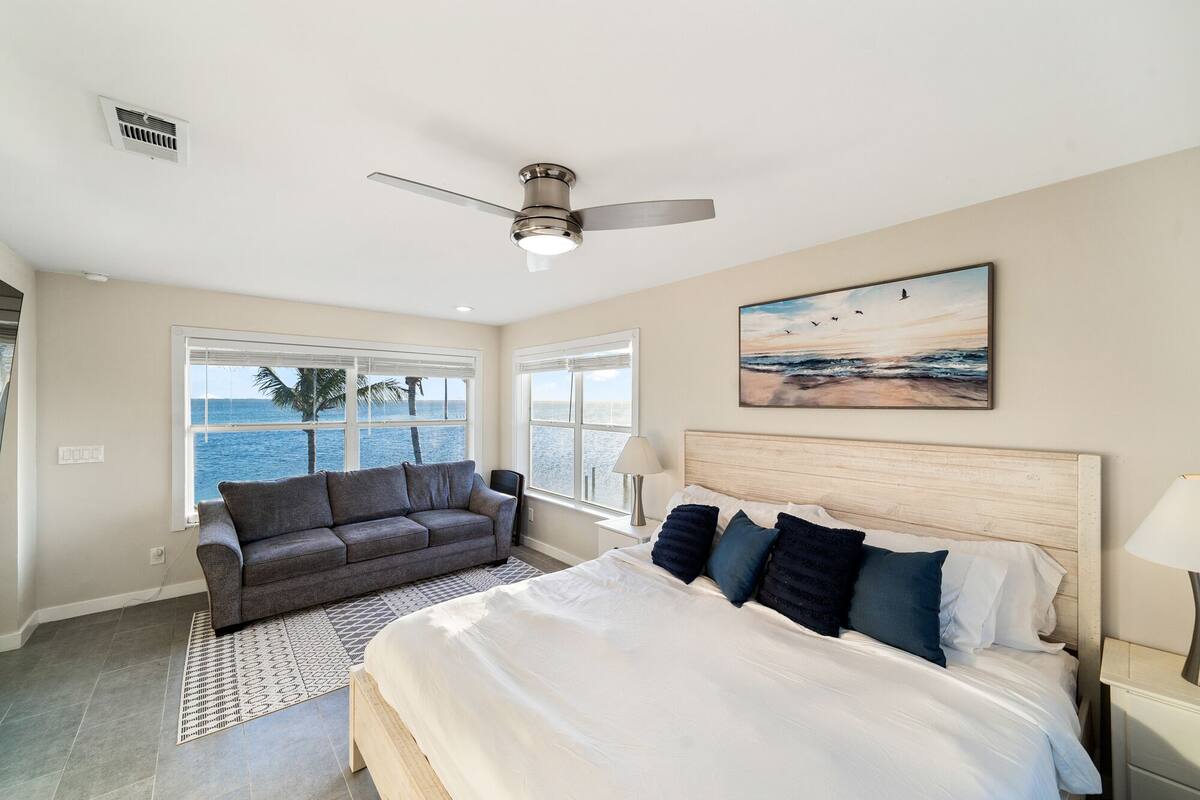 A bedroom with a large bed, a gray sofa, a ceiling fan, and a painting of birds over the ocean.