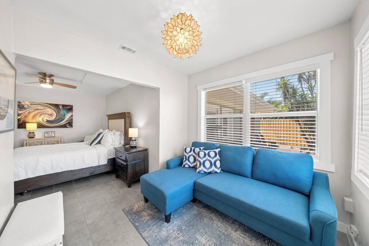 A modern bedroom with a bed, a blue sofa, a ceiling fan, and a large window with blinds.