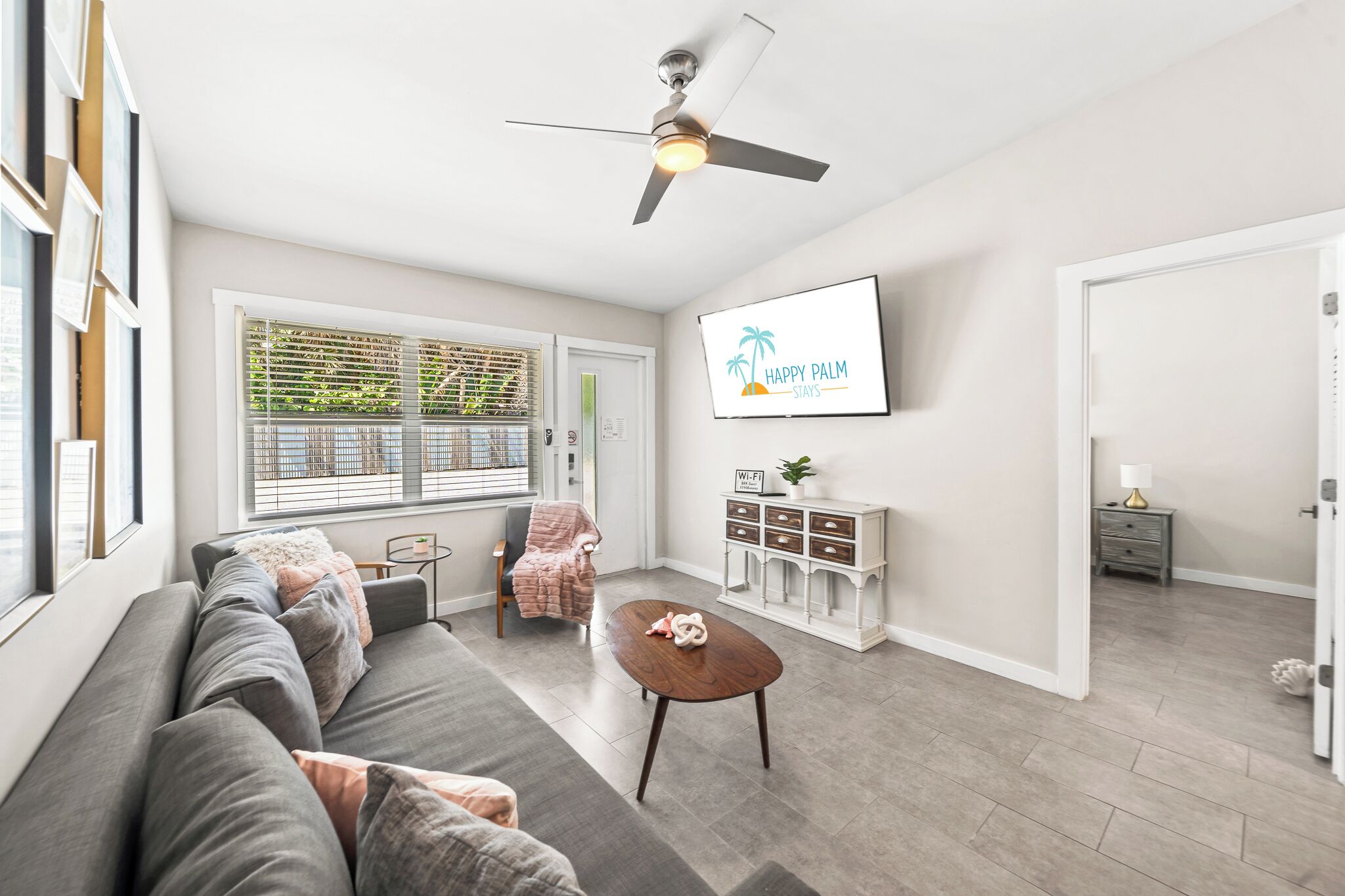 A modern living room with a grey sofa, a wooden coffee table, a ceiling fan, a large window with blinds, a white cabinet, and a framed sign on the wall.