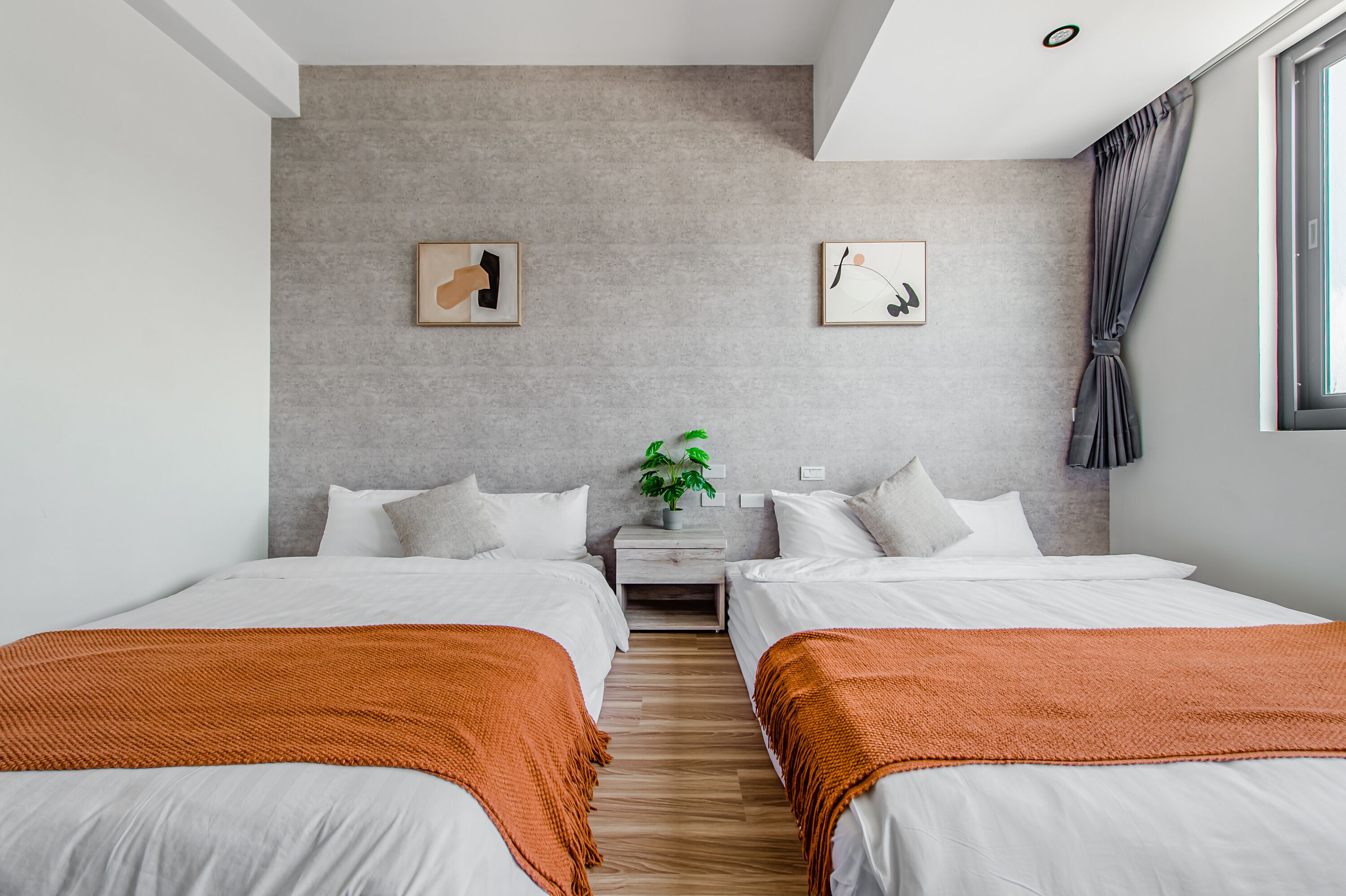 A modern hotel room with two beds, a grey textured wall, and a small bedside table with a plant.