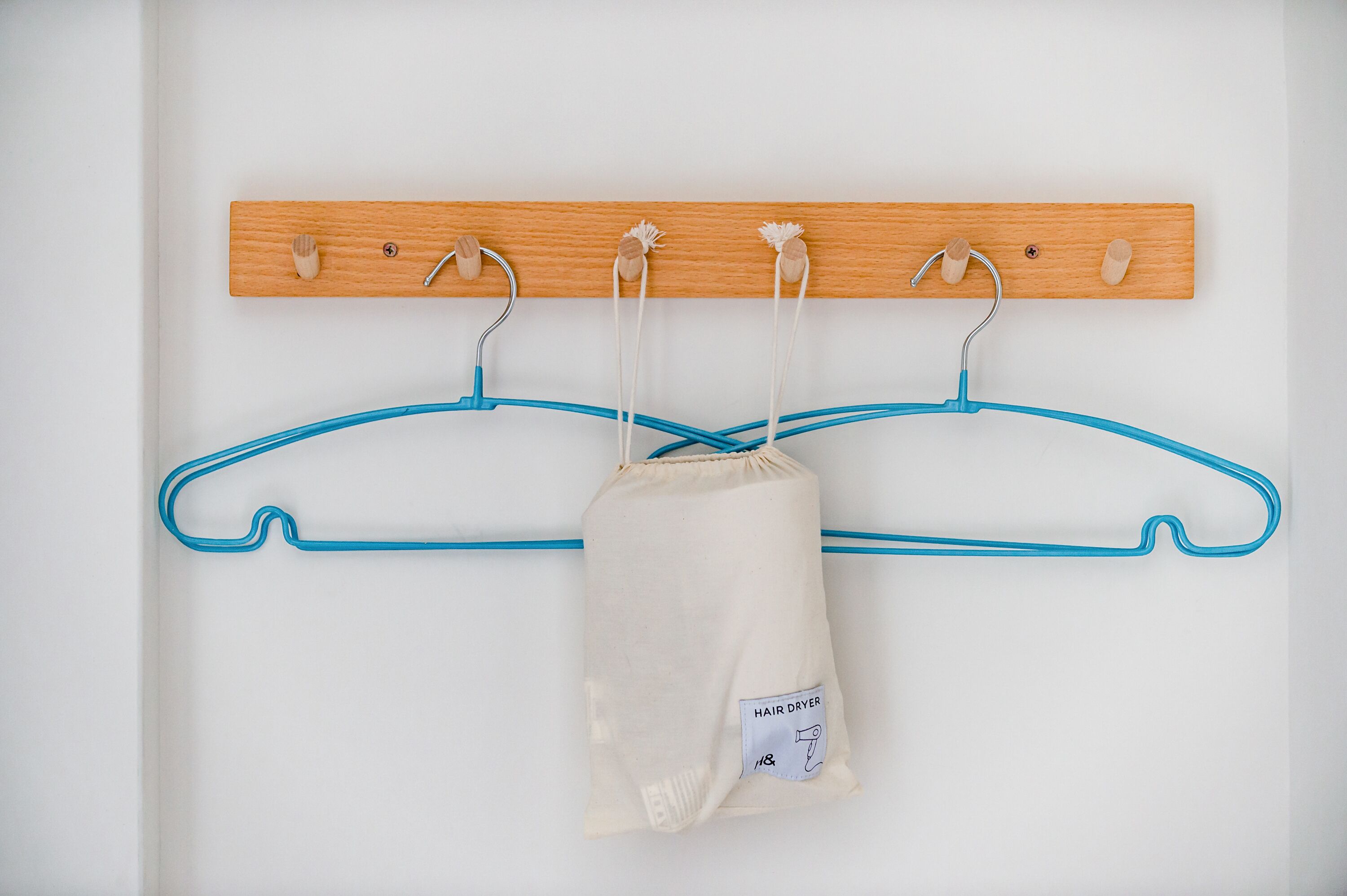 A wooden coat rack with four hooks, two blue hangers, and a beige garment bag labeled "HAIR DRYER".