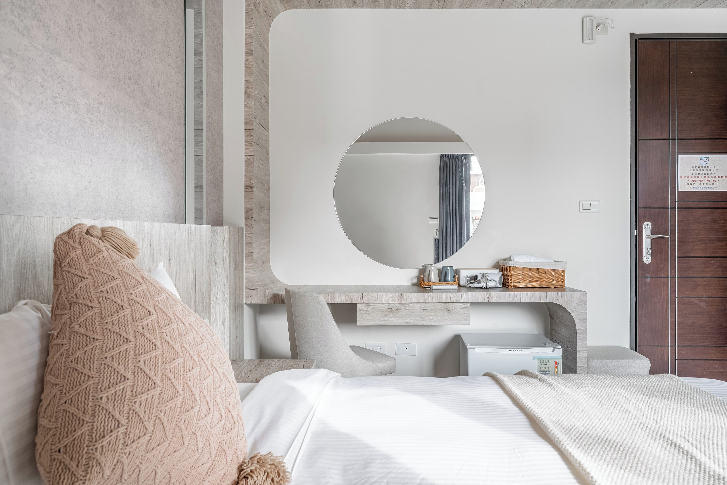 A modern bedroom with a large bed, a round mirror, a wooden dresser, and a door with a sign.