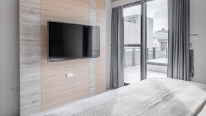 Et moderne hotelværelse med et fladskærms-tv monteret på en væg af træpaneler, en balkon med grå gardiner og en seng med et hvidt, struktureret tæppe.