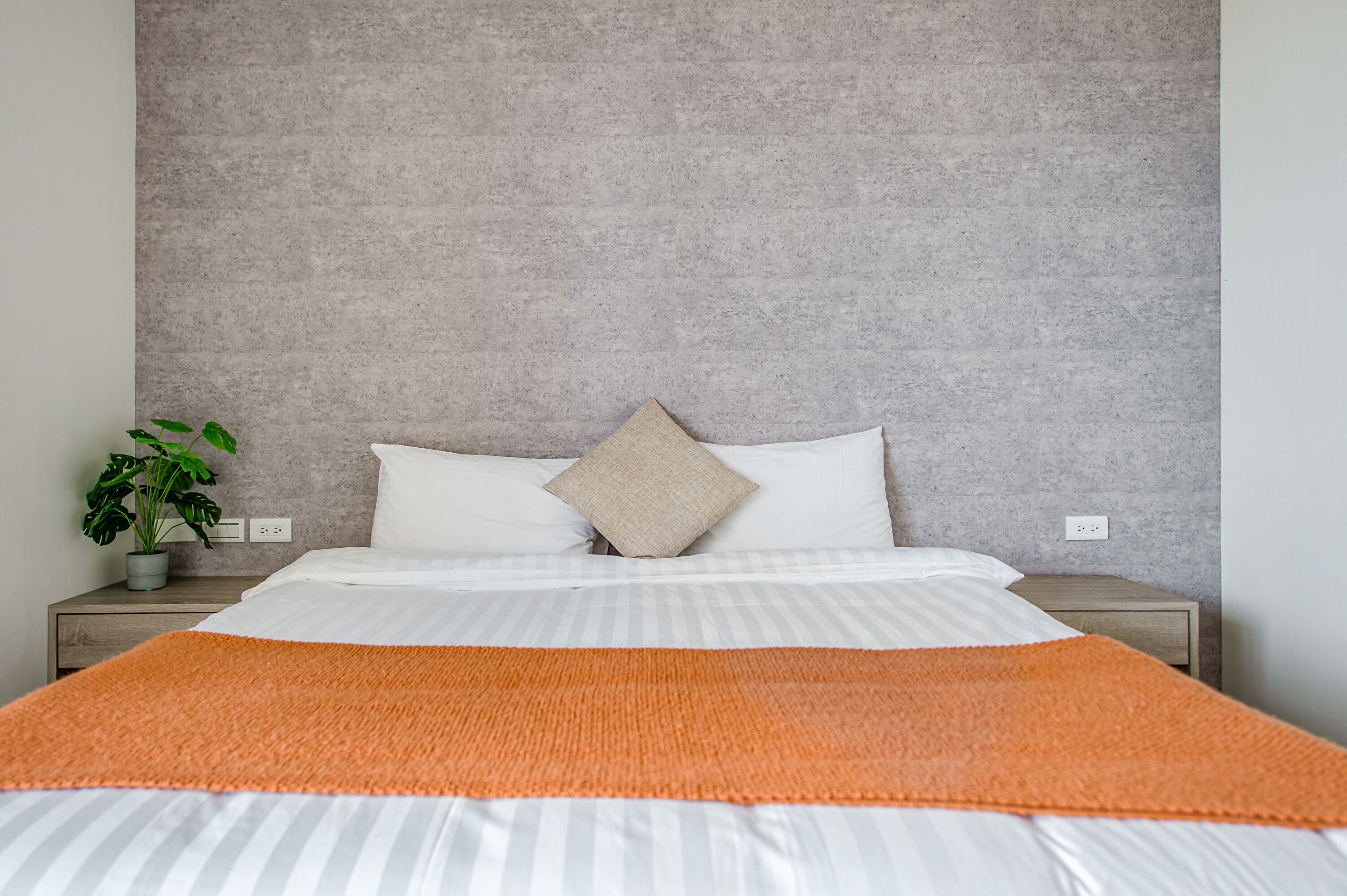 A neatly made bed with a striped white and orange blanket, a grey textured headboard, and a single potted plant on the nightstand.