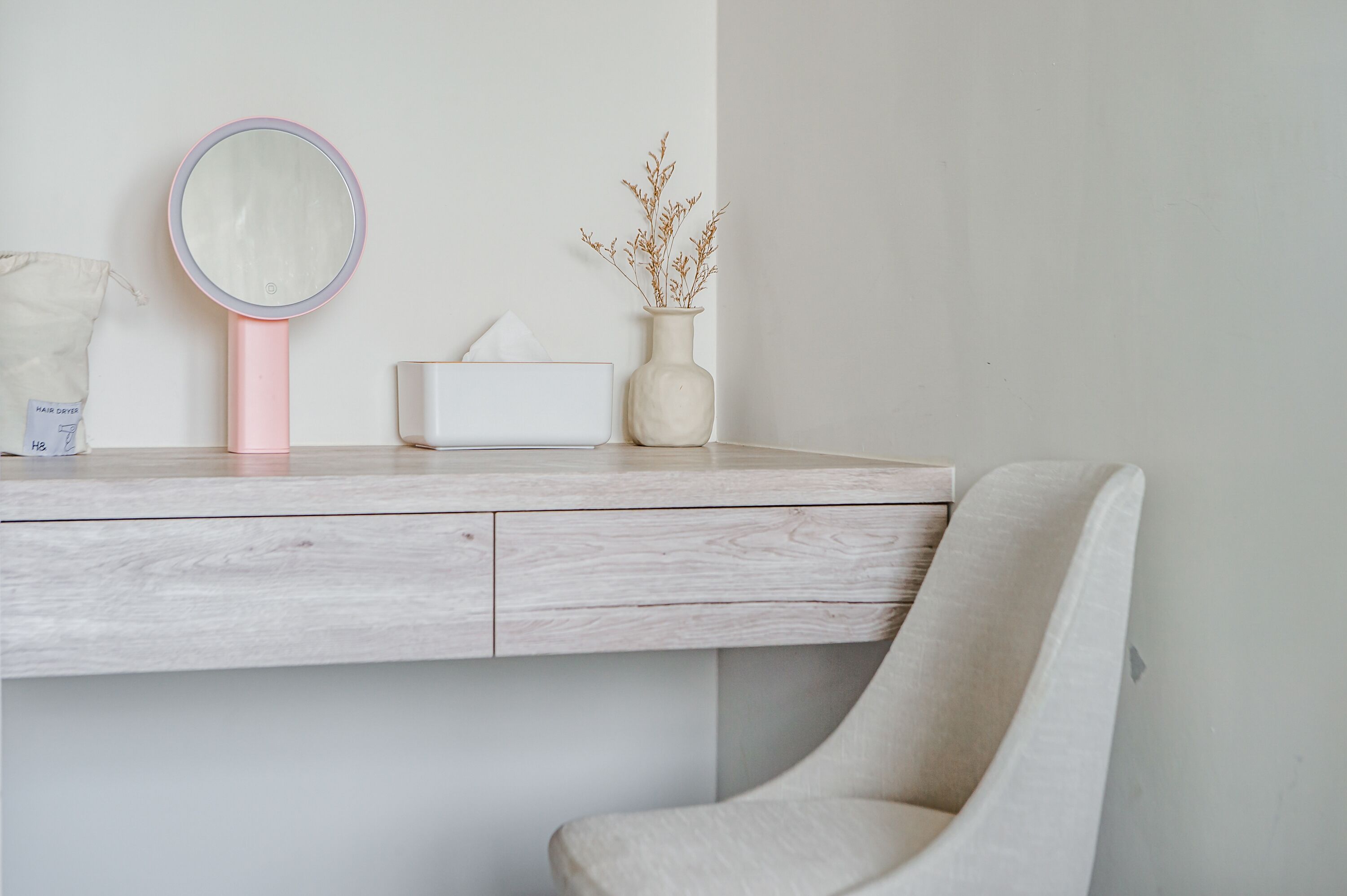 Un tocador minimalista con superficie de madera, espejo rosa, caja de pañuelos blancos y un florero con plantas secas.