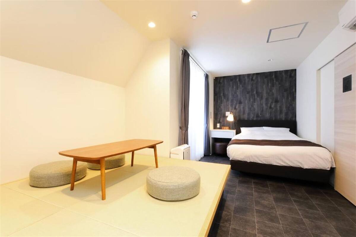 Une chambre d’hôtel moderne équipée d’un lit, d’une table de chevet, d’une lampe et d’un petit coin salon avec une table et un pouf.