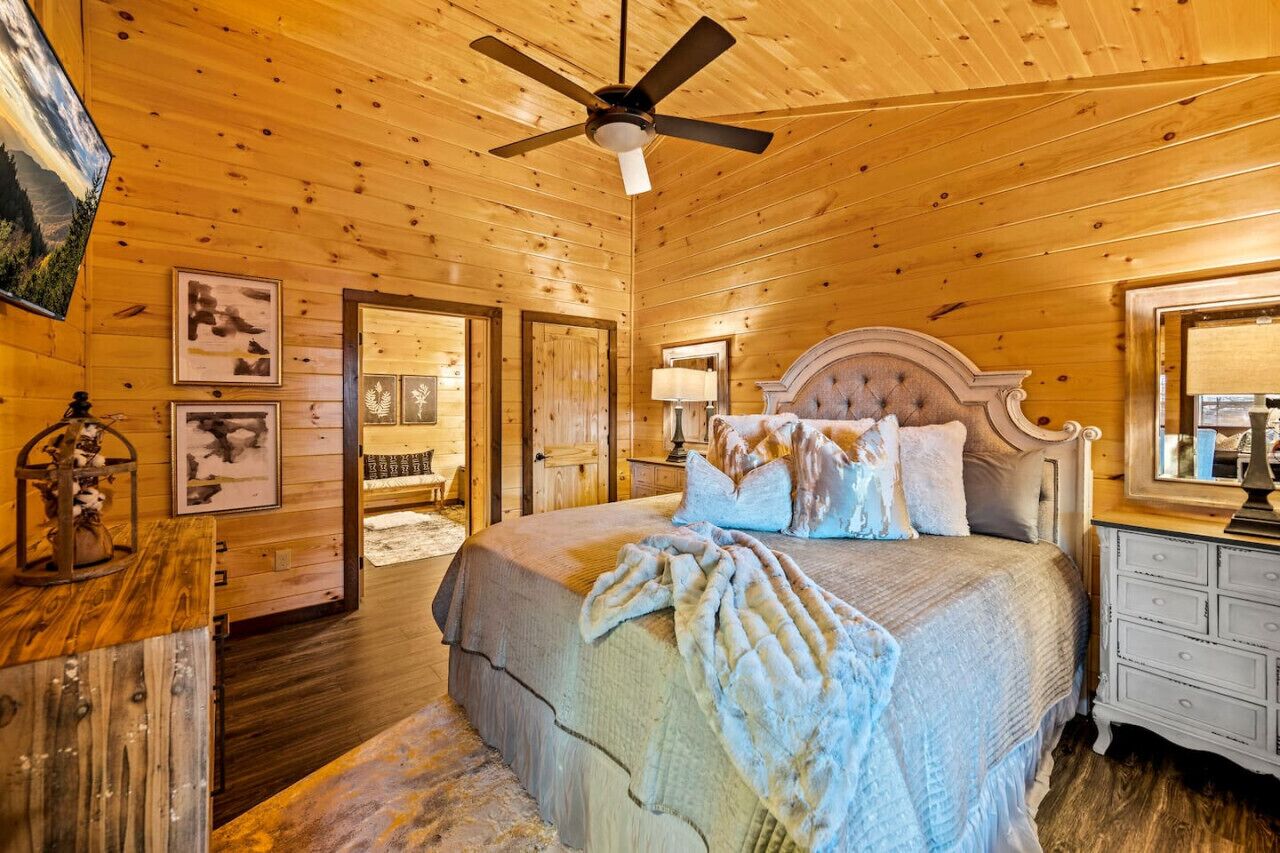 A wooden-paneled bedroom with a large bed, a ceiling fan, and framed artwork on the walls.