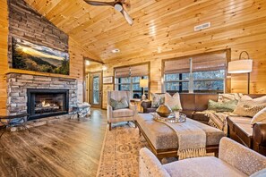 A cozy cabin interior with a stone fireplace, wooden ceiling, and large windows.