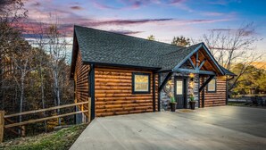 Une cabane en rondins, avec des fondations en pierre, une allée en béton et une vue sur la forêt environnante.