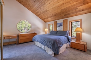 A bedroom with a wooden ceiling, a round window, a bed with blue bedding, two bedside tables with lamps, and a dresser.