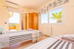 A bedroom with two beds, a window with a view of palm trees, an air conditioning unit, and a door leading to an outdoor balcony.
