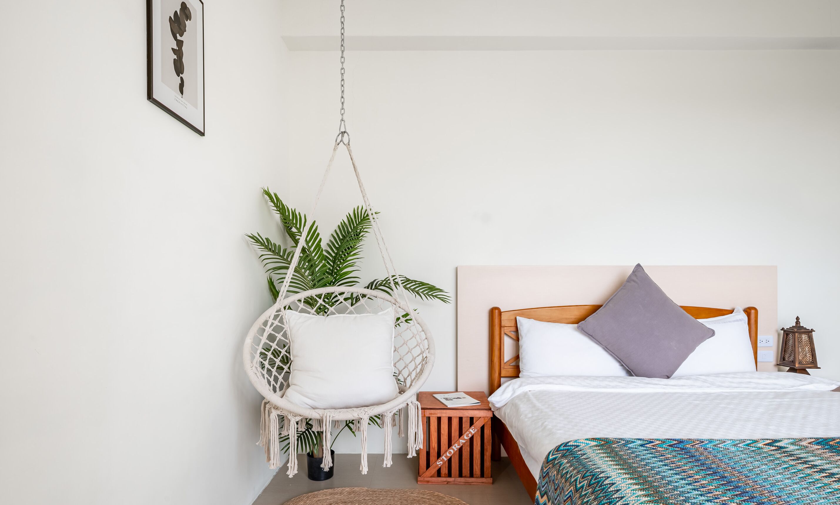 A bedroom with a bed, a hanging chair, a small wooden side table, and a potted plant.