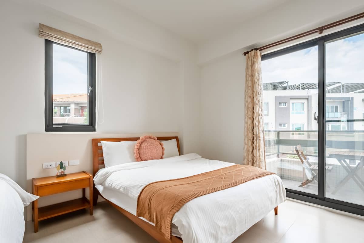 Ein Schlafzimmer mit großem Fenster, einem Bett mit weißer Bettwäsche, einem Nachttisch aus Holz und Blick auf Gebäude im Freien.