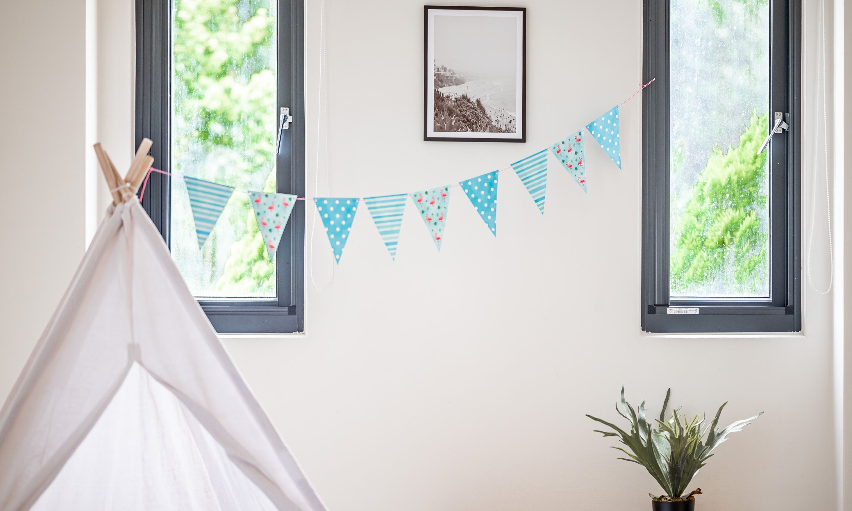 Une pièce avec deux fenêtres, une tente blanche, une guirlande de drapeaux triangulaires bleus et blancs, un tableau encadré accroché au mur et une plante en pot.