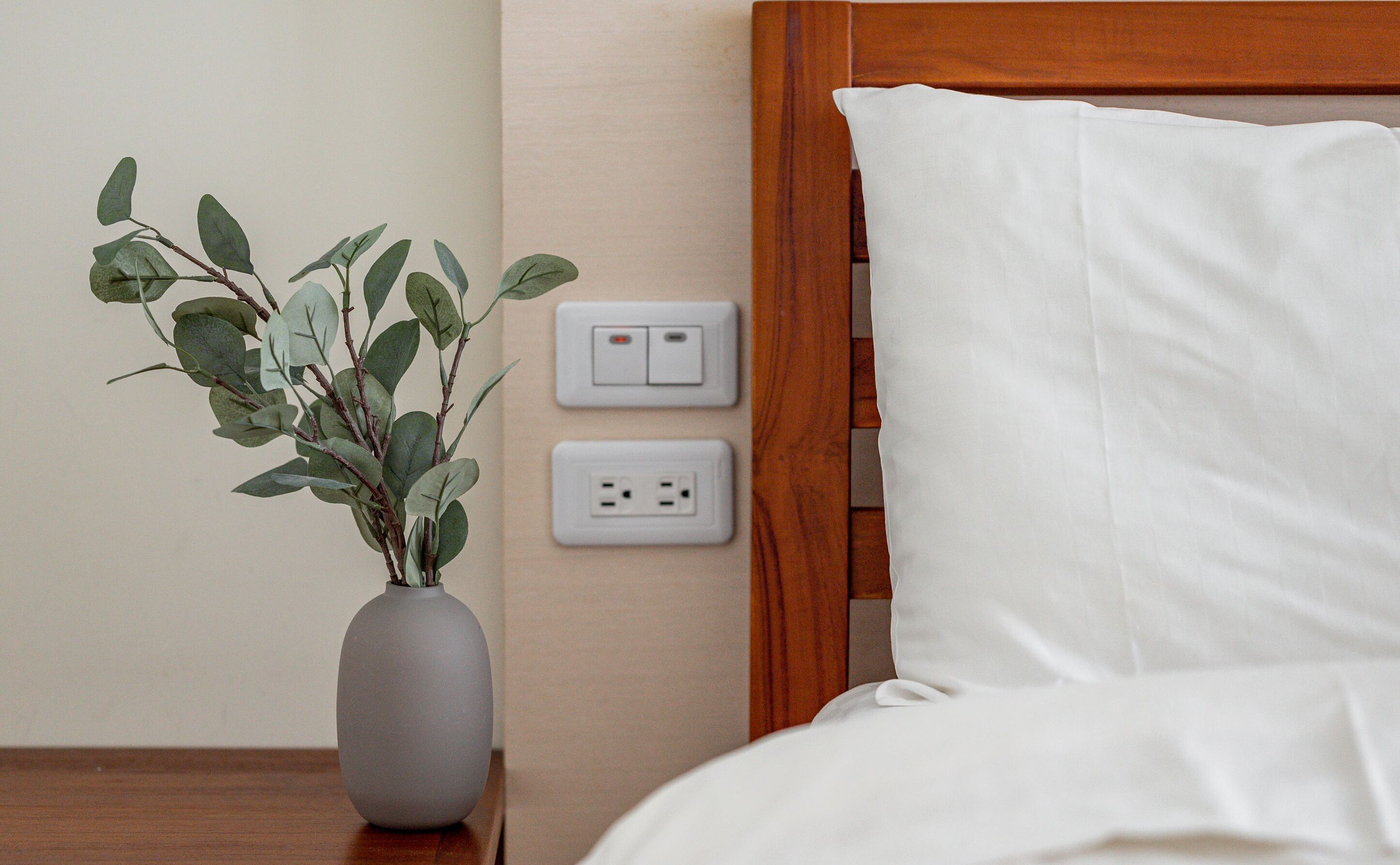 A bed with white sheets, a grey vase with green leaves, and two electrical outlets on the wall.