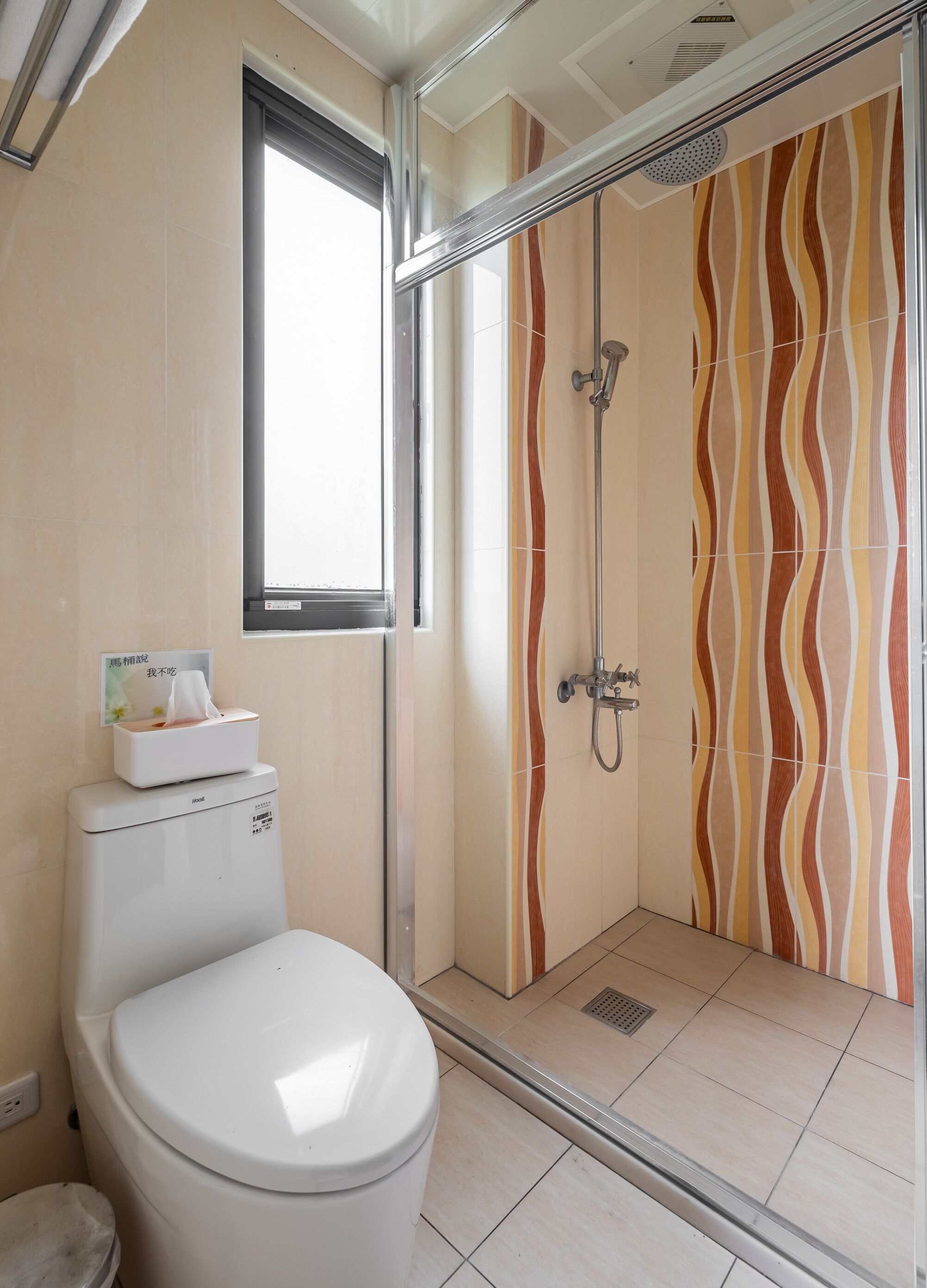Une salle de bain avec des toilettes, une douche dont les carreaux sont disposés en motifs ondulés, et une fenêtre.