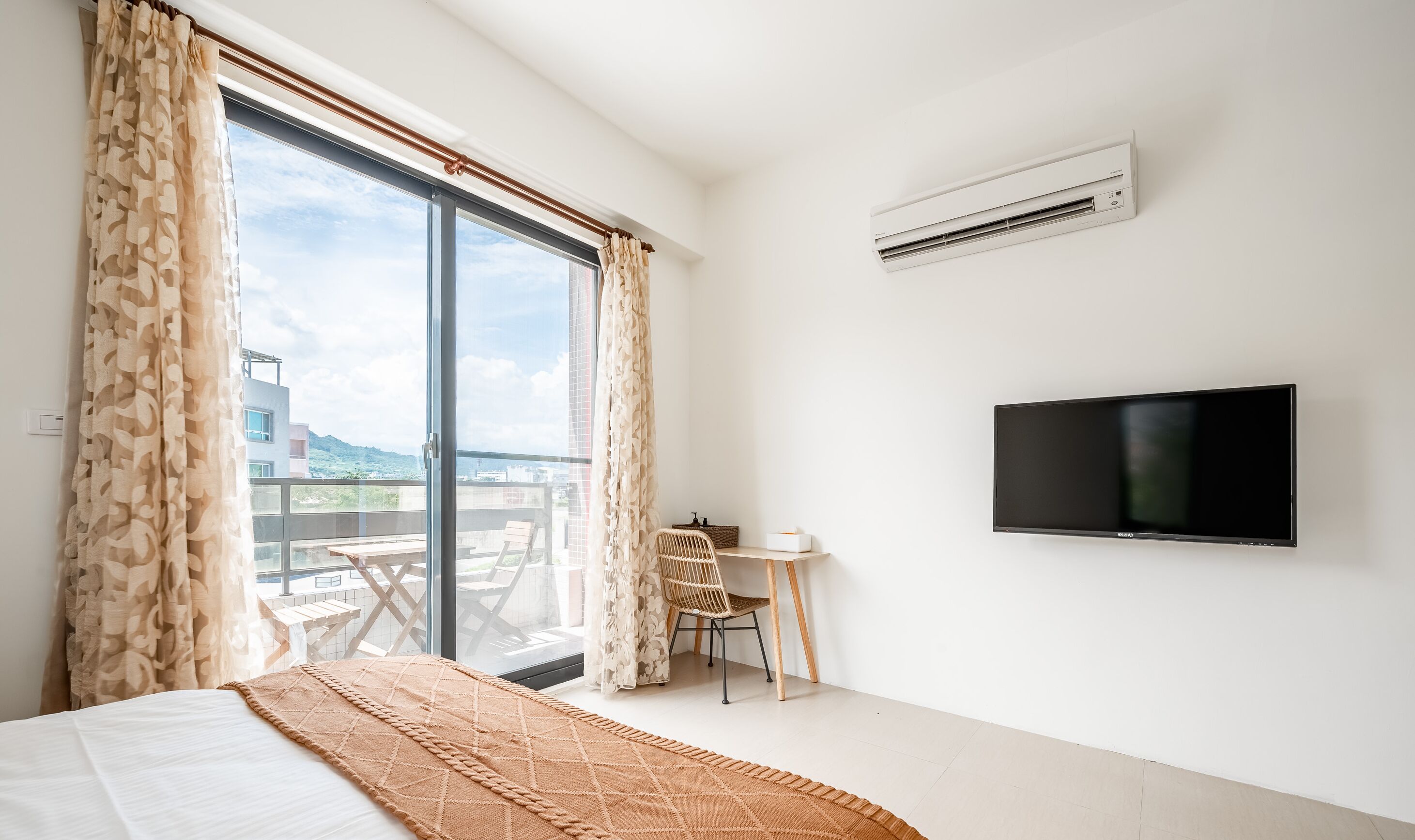 A hotel room with a bed, a TV mounted on the wall, a small table with a chair, and a balcony with a table and chairs.
