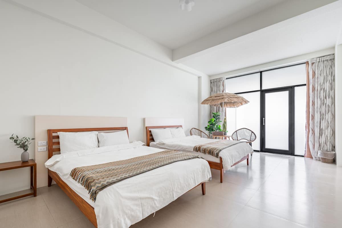 Ein geräumiges Schlafzimmer mit zwei Betten, einem großen Fenster mit Vorhängen, einer Topfpflanze und einem gewebten Sonnenschirm.