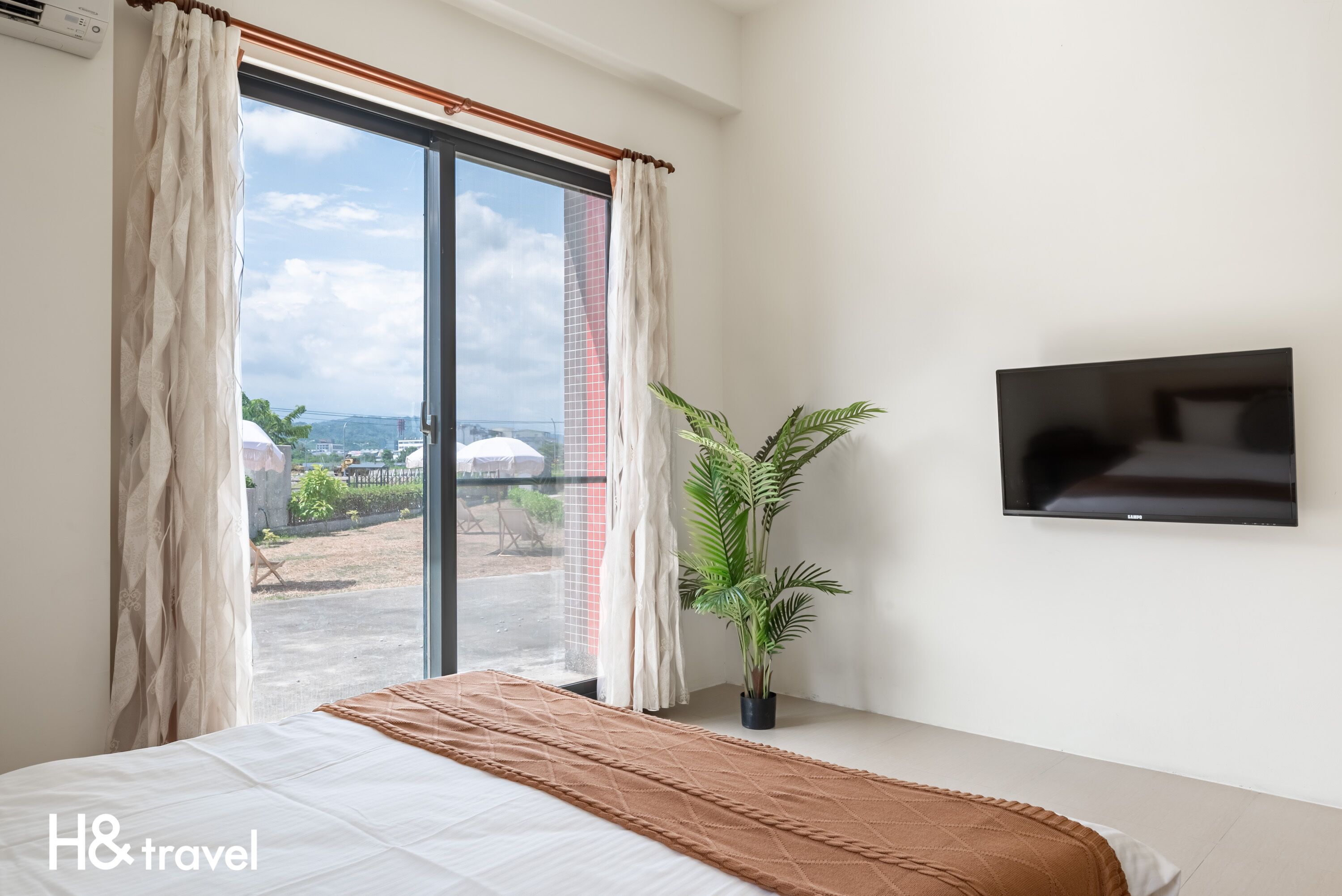 A hotel room with a bed, a potted plant, a TV, and a large window with curtains.