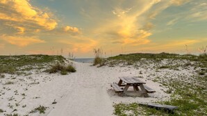 Uma trilha de areia que leva a uma mesa de piquenique de madeira com um banco, cercada por vegetação rala.
