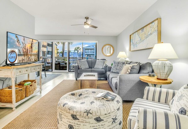 A living room with a large flat-screen TV, a ceiling fan, a sofa, a coffee table, and a side table with a lamp.