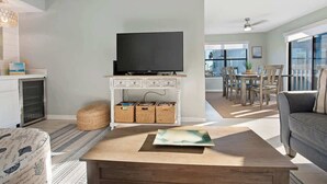 Uma sala de estar moderna com uma TV de tela plana em um armário branco, uma mesa de centro de madeira, um sofá cinza e uma área de jantar com cadeiras e mesa de madeira.