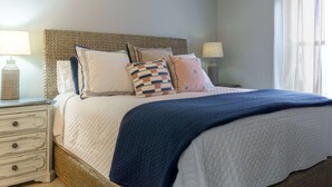 Cama bem arrumada com cobre acolchoado, cabeceira e vários travesseiros. Tem uma mesa de cabeceira com abajur e um vaso decorativo.