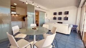 Una sala de estar moderna con una mesa de comedor de vidrio rodeada por sillas blancas, un sofá blanco y una cocina al fondo.