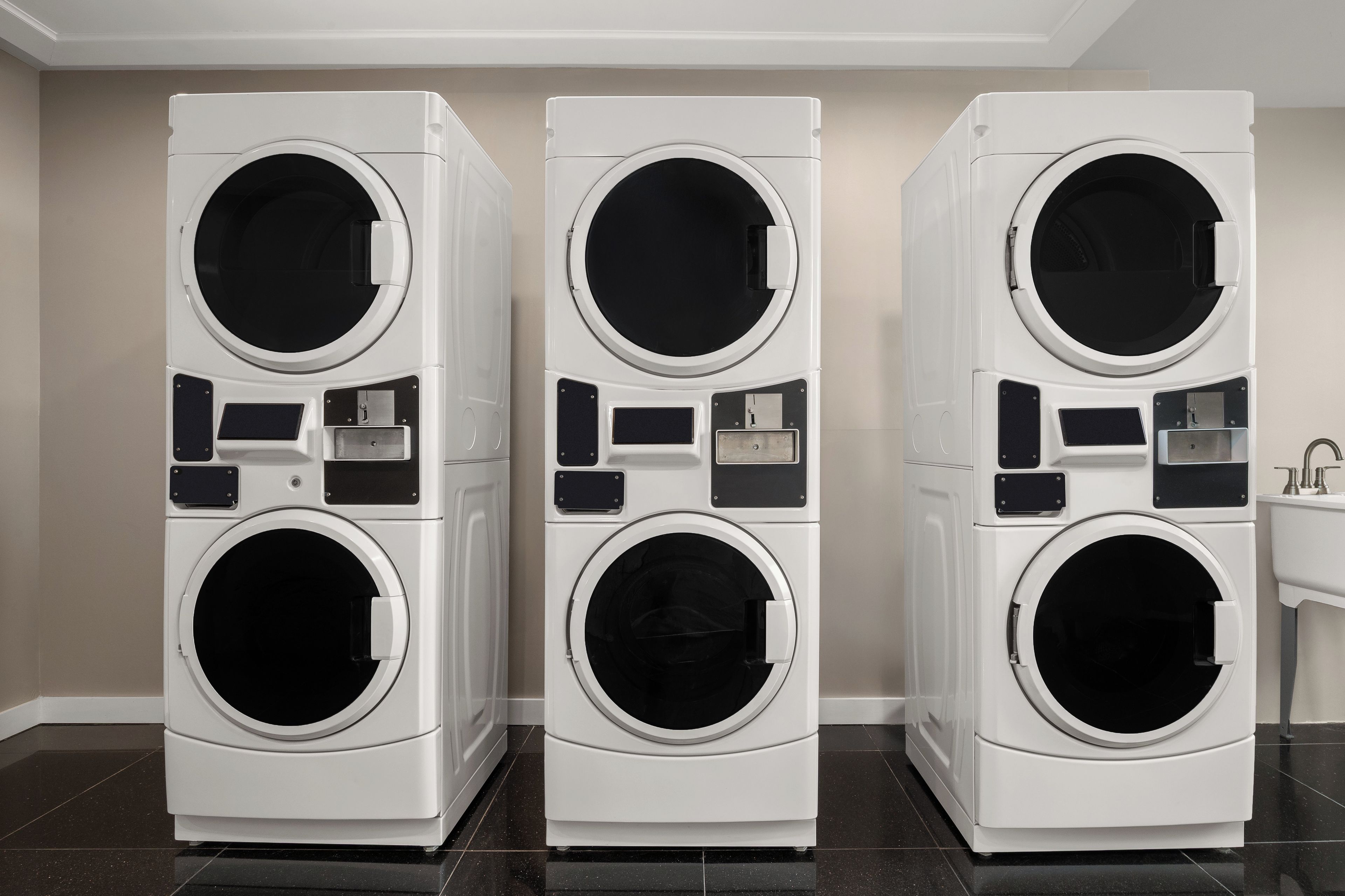 Three stacked front-loading washing machines in a room with a tiled floor and a sink on the right.