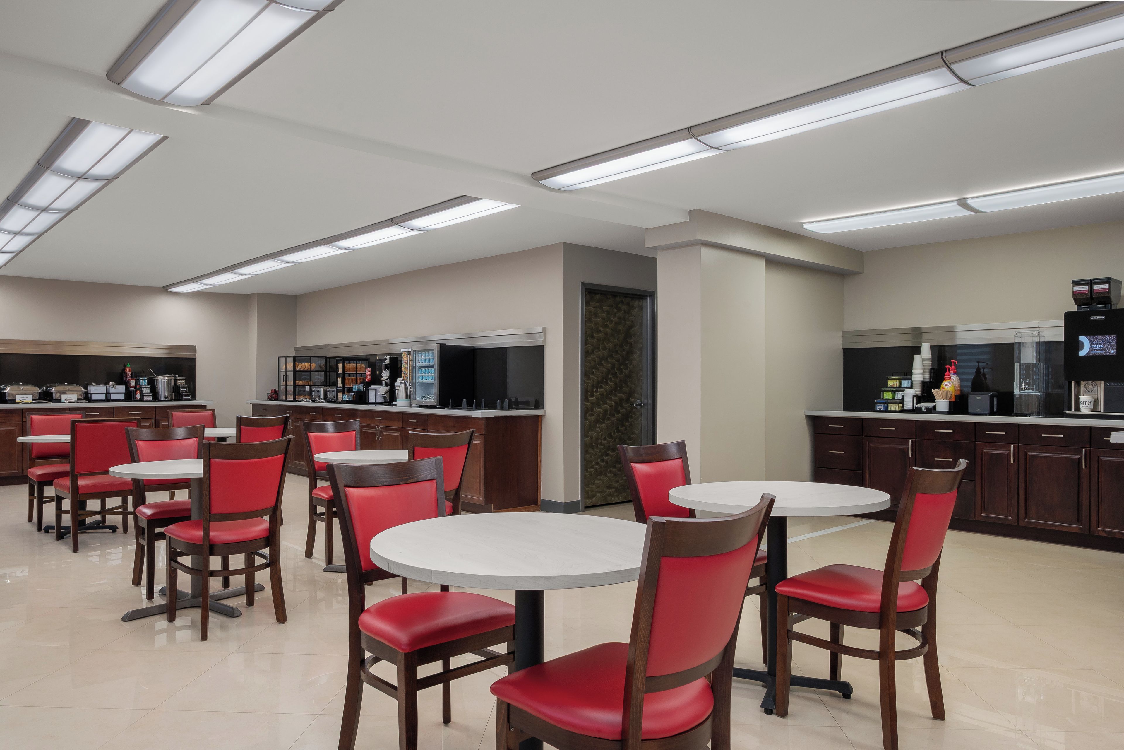 A modern restaurant with round tables and red chairs, a bar area, and a kitchen in the background.