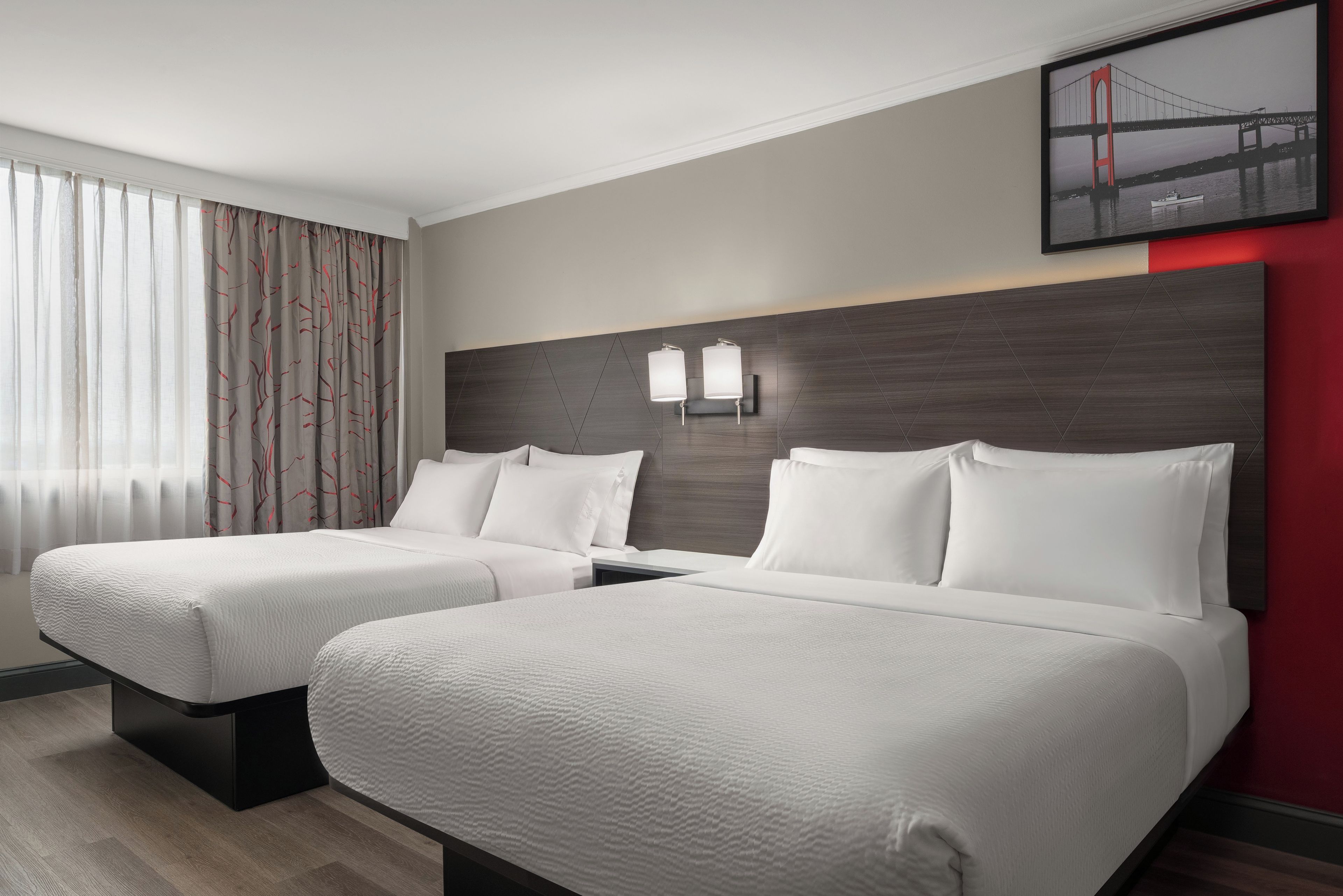 A hotel room with two beds, a headboard with wall-mounted lights, a window with sheer curtains, and a framed picture of a bridge.