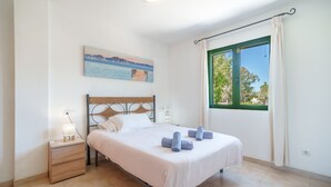 A bedroom with a bed, a nightstand, a painting, a window with curtains, and a view of trees and a building. - Sunwing Beach by Interhome (Port d'Alcúdia)