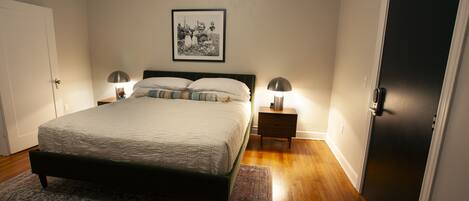 A bedroom with a bed, two bedside tables, a nightstand, a framed picture on the wall, and a door.