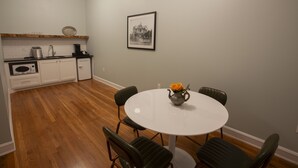 A compact kitchen with white cabinets, a wooden countertop, and a coffee maker. A dining table with four chairs and a vase of flowers is in the foreground. - Greyhound Inn - Deluxe King Suite (Tyler)