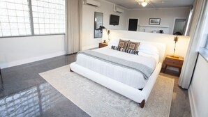 A modern bedroom with a large bed, a bedside table, a nightstand, a TV, and a window with curtains. - Greyhound Inn - Deluxe King Suite (Tyler)