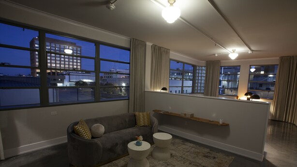 A modern living room with a grey sofa, two round side tables, a large window with a view of a building and a body of water, and a ceiling light fixture. - Greyhound Inn - Deluxe King Suite (Tyler)