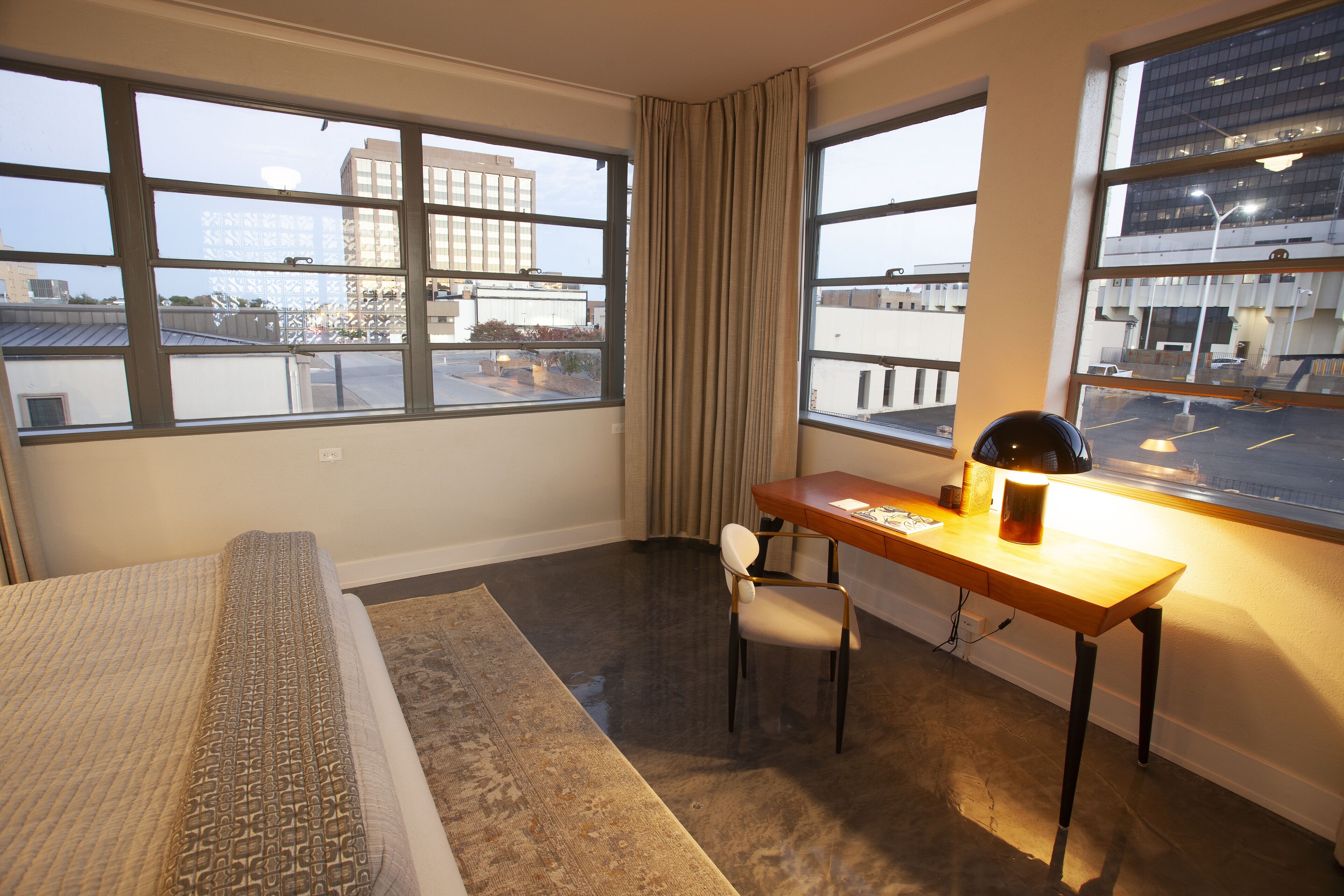 A hotel room with a bed, a desk, a chair, and large windows offering a view of the cityscape.