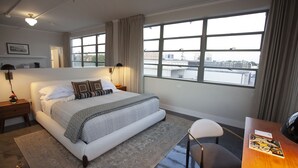 A modern hotel room with a large bed, a desk, and a view of the city through large windows. - Greyhound Inn - Deluxe King Suite (Tyler)