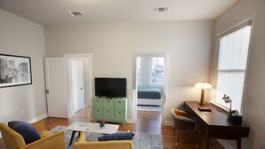 Un espacio habitable pequeño con un sofá, una mesa de centro, un televisor sobre un mueble verde, un escritorio con una lámpara y una ventana con persianas.