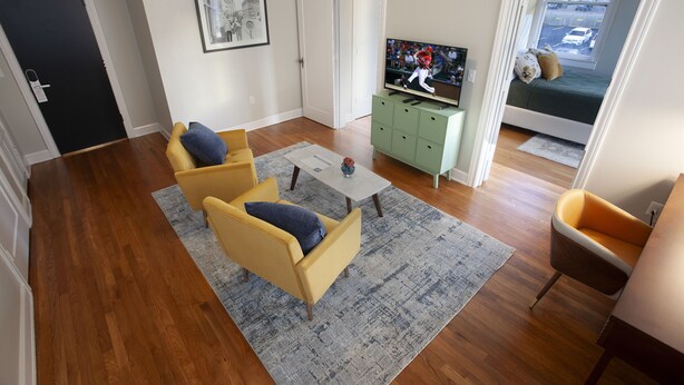 Una sala de estar con dos sillones amarillos, un aparador verde, un televisor y vista a la calle a través de una puerta.
