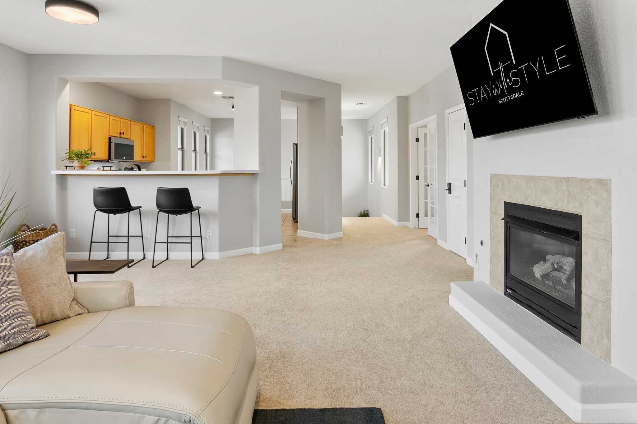 Una sala de estar moderna con un sofá color beige, una chimenea y una zona de cocina con armarios amarillos y una barra.