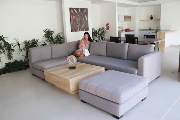 A modern living room with a grey sectional sofa, a wooden coffee table, and a small ottoman. A person is seated on the sofa, talking on the phone. The room has a minimalist design with a kitchen area visible in the background.