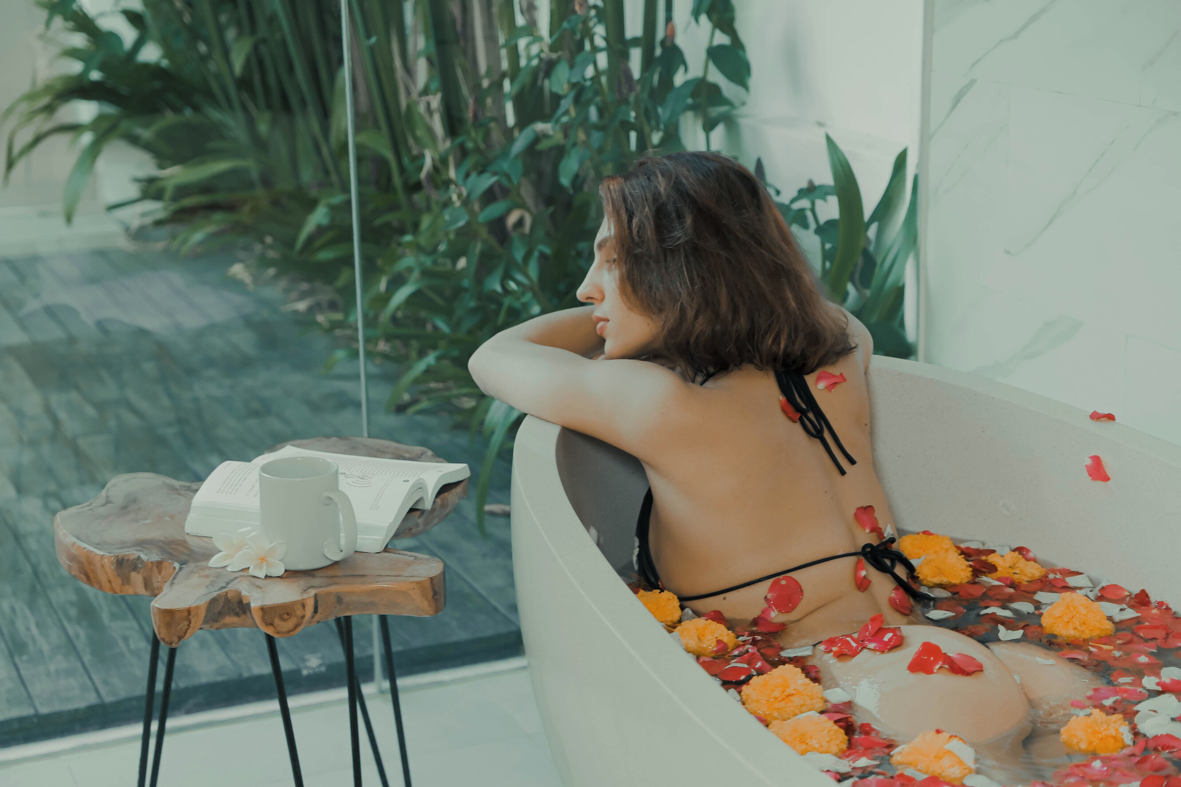 A bathtub filled with water and rose petals, a book, and a mug on a wooden stool.