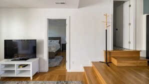 Un salon moderne avec un téléviseur à écran plat posé sur un meuble blanc, un parquet en bois, un escalier et une porte-fenêtre donnant sur une autre pièce.