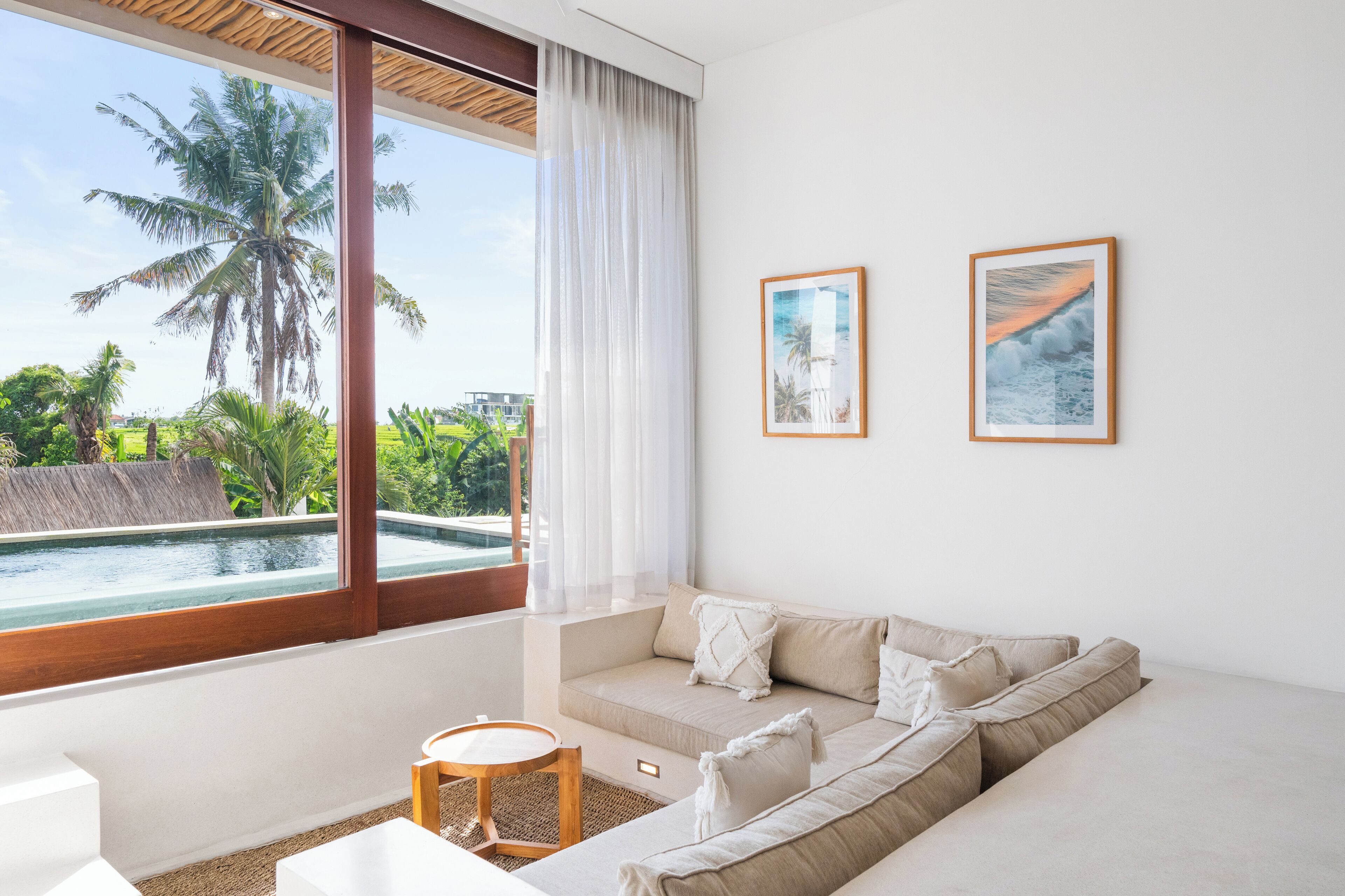 A modern room with a large window, a beige sofa, a small round table, and two framed pictures on the wall.