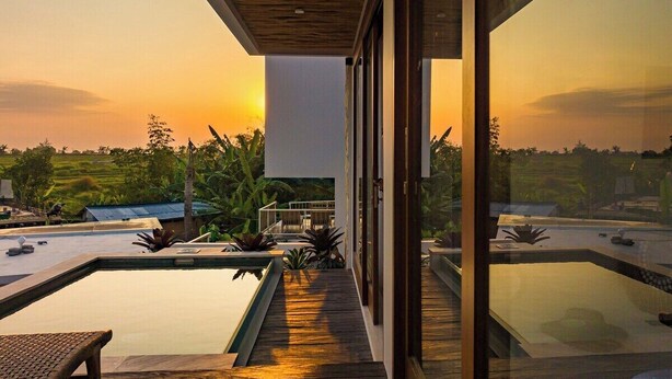 Un área de piscina moderna al aire libre con muelle de madera, paredes de vidrio y vista al atardecer sobre un paisaje.