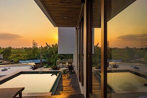 A modern outdoor pool area with a wooden deck, glass walls, and a view of the sunset over a landscape. - Pagi Villas by Betterplace (Canggu)