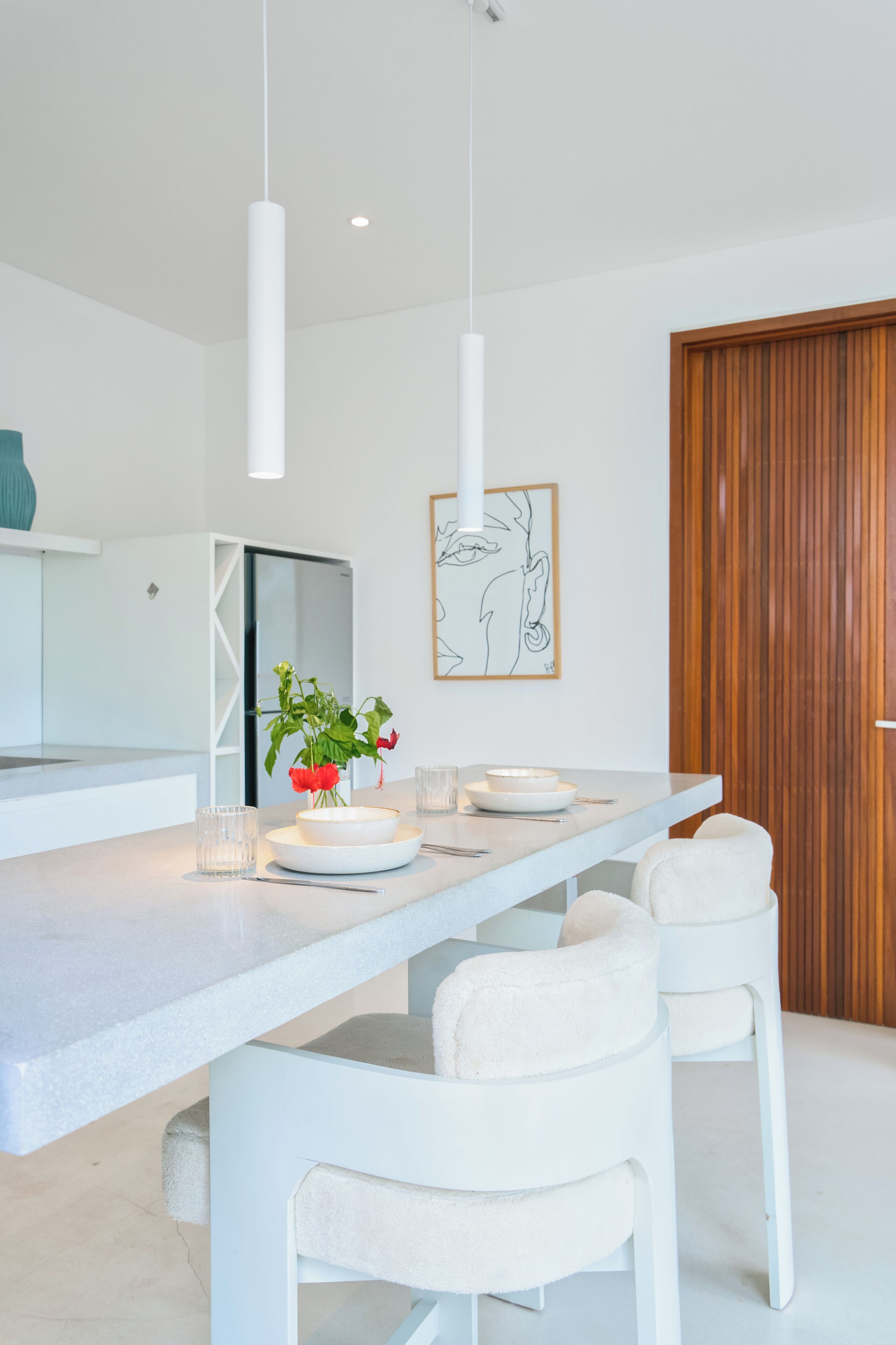 A modern kitchen with white countertops, a central island with seating, pendant lighting, and a wooden door.